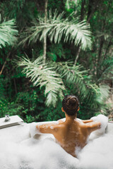 Woman relaxing in bath tub full of foam outdoors with jungle view. View from behind. Unrecognizable person. Beauty spa treatment, leisure time. - 386190171