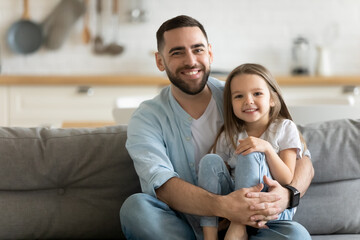 Portrait smiling father and little daughter hugging, looking at camera, sitting on cozy couch at...