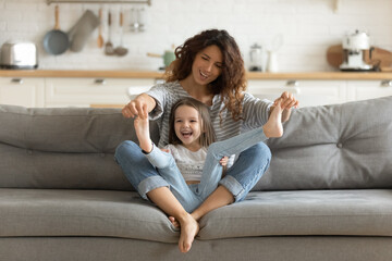 Overjoyed young mother and little daughter having fun, caring mum holding cute preschool girl legs, enjoying funny activity, sitting on cozy couch at home, family spending leisure time together - Powered by Adobe