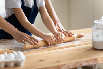 Close up caring mother and little daughter wearing aprons rolling out dough together, working with rolling pin, standing at wooden countertop in kitchen, family cooking baking together - Powered by Adobe