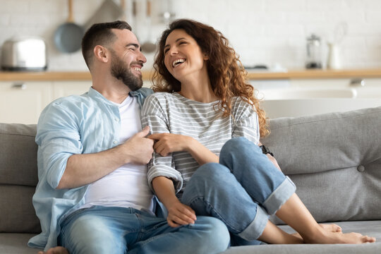 Happy Married Young Couple Hugging, Sitting On Cozy Couch Together, Overjoyed Laughing Woman And Man Having Fun, Enjoying Leisure Time, Relaxing On Sofa In Living Room At Home, Good Relationship