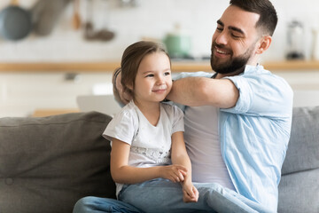 Caring smiling father brushing combing adorable preschool daughter hair, helping with hairstyle, kid morning routine, cute little girl sitting on dad laps, sitting on cozy couch at home