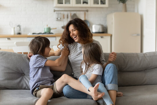 Overjoyed Young Mother Having Fun With Two Kids At Home, Laughing Happy Mum, Adorable Little Daughter And Son Playing Funny Active Game, Tickling, Sitting On Cozy Couch In Living Room