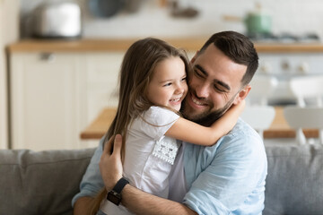 Close up smiling loving young father hugging adorable little daughter, enjoying tender moment, spending weekend together, sitting on cozy couch at home, good family relationship between dad and child - obrazy, fototapety, plakaty