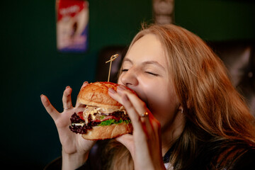 Portrait of a young hungry woman sitting in the street fast junk food restaurant with open mouth bite and eating enjoying a fresh tasty burger. Image with copy space.
