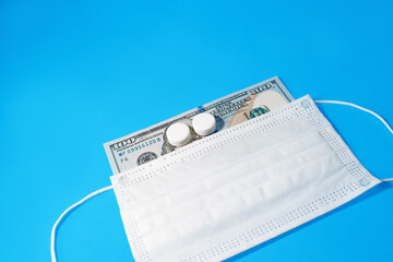 One hundred dollars under a medical face mask on a blue background, copy space. The pills lie in front of Benjamin Franklin. Global recession, covid-19, medical and health spending.