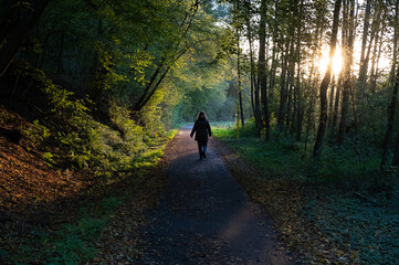 Woman walking on a road through a forest, the low sun shining through the trees.