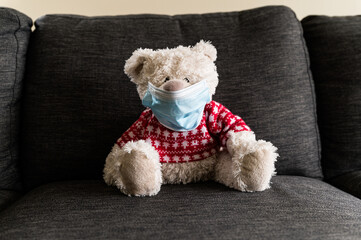 Cute teddy bear wearing a protective mask sitting on a sofa.