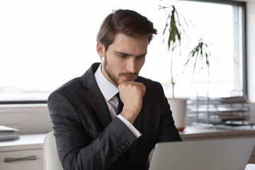 Head shot focused young smart businessman in formal suit looking at computer screen, solving difficult problem or analyzing project risks alone at workplace, corporate employee casual office workday.