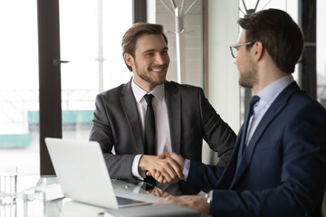Smiling two young businessman in formal suits shaking hands, making agreement after online project presentation on computer. Happy manager thanking employee for presenting development strategy.
