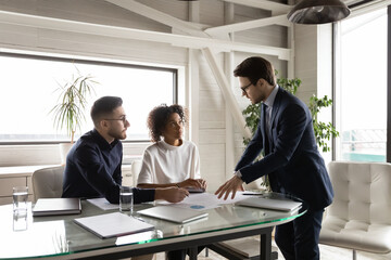 Concentrated young mixed race employees listening to skilled 30s ceo executive manager, explaining company sales strategy at briefing meeting in modern office, multiracial business people negotiating.