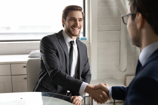 Smiling young male job seeker shaking hands with employer hr manager, making good first impression after successful job interview in office. Happy two businessmen celebrating making agreement.