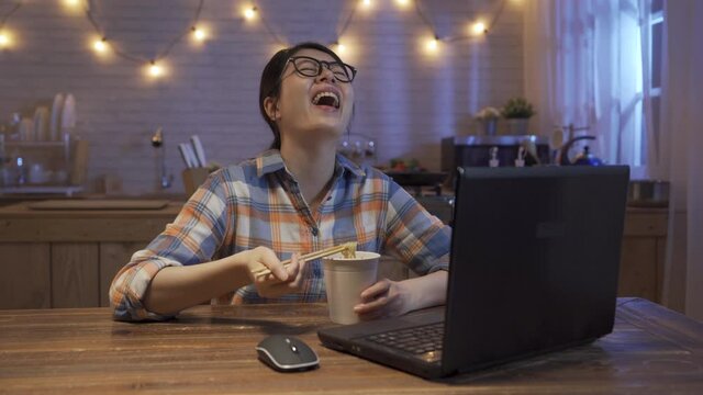 relax young asian japanese girl eating instant noodles and laughing cheerfully while watching online funny video on laptop computer