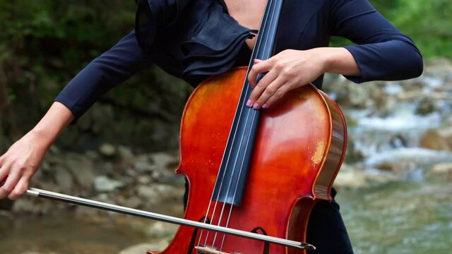 Woman plays the cello outdoors. Female musician playing the instrument on natural river background in summer. Musical equipment in womans' hands.