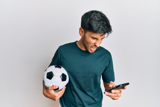 Young handsome man holding football ball looking at smartphone bet app angry and mad screaming frustrated and furious, shouting with anger. rage and aggressive concept.