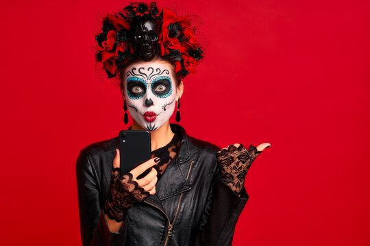 Girl with sugar skull makeup with a wreath of flowers on her head and skull, wearth lace gloves and leather jacket, shocked with news from her phone isolated on red background