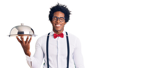 Handsome african american man with afro hair wearing waiter uniform holding silver tray looking positive and happy standing and smiling with a confident smile showing teeth