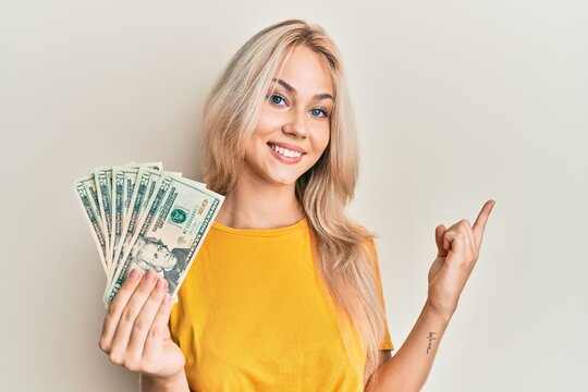Beautiful caucasian blonde girl holding 20 dollars banknotes smiling happy pointing with hand and finger to the side