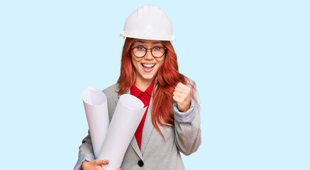 Young redhead architect woman wearing safety helmet holding blueprints screaming proud, celebrating victory and success very excited with raised arms