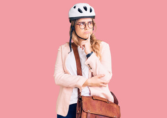 Young caucasian woman wearing bike helmet and leather bag serious face thinking about question with hand on chin, thoughtful about confusing idea