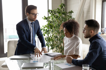 Serious 30s male team leader in formal suit and eyeglasses negotiating marketing research results with focused african and arabian ethnicity colleagues, holding brainstorming meeting in office.