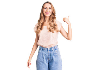 Young beautiful caucasian woman with blond hair wearing casual clothes smiling with happy face looking and pointing to the side with thumb up.
