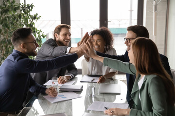 Overjoyed young mixed race business people sitting at table, joining hands in air, showing group...