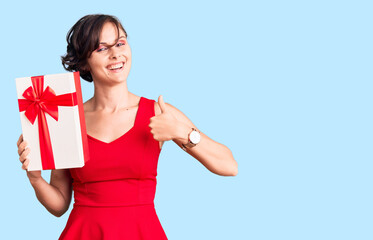 Beautiful young woman with short hair holding gift smiling happy and positive, thumb up doing excellent and approval sign