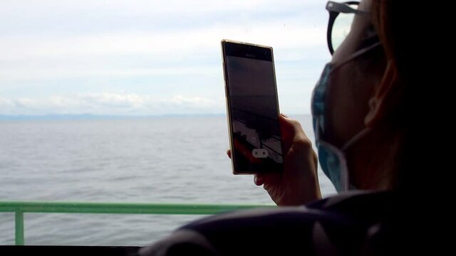 asian woman using mobile smart phone taking video on ferry boat. female wearing medical face mask when travel during covid19 pandemic outbreak. new normal.