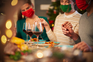 Group of friends in masks praying over Christmas Thanksgiving table at home