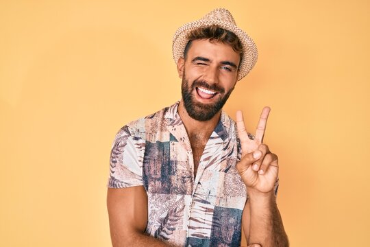 Young hispanic man wearing summer hat smiling with happy face winking at the camera doing victory sign. number two.