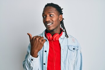 African american man with braids listening to music using headphones smiling with happy face looking and pointing to the side with thumb up.