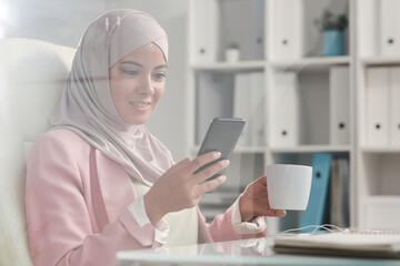 Happy restful Muslim businesswoman in hijab scrolling in smartphone at break