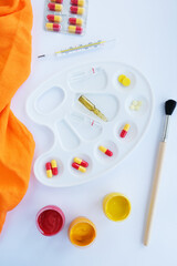 On a white background, a palette for paints and in it are pills, ampoules and a thermometer, next to it there are paints with a brush.

