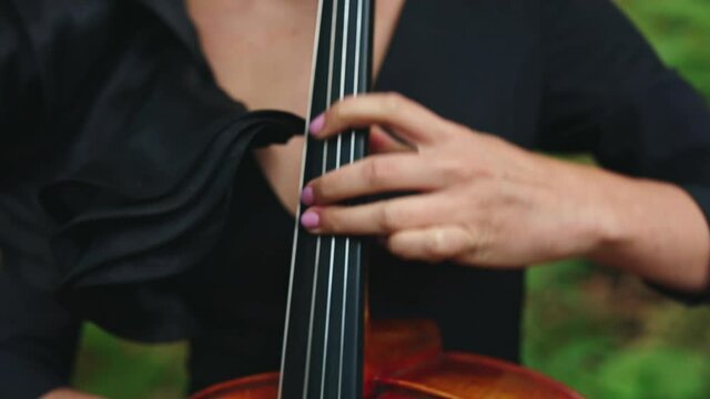 Cello playing. Woman's fingers perform music on strings of a cello. Musical instrument in female's hands outdoors. Close-up.