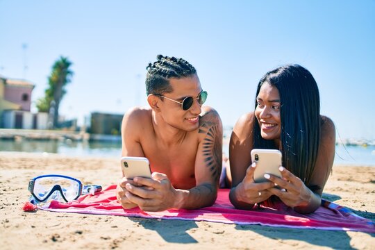 Young latin couple using smartphone lying on the sand at the beach.