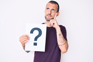 Young handsome man with tattoo holding question mark serious face thinking about question with hand on chin, thoughtful about confusing idea