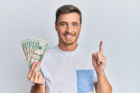 Handsome caucasian man holding 20 dollars banknote smiling with an idea or question pointing finger with happy face, number one