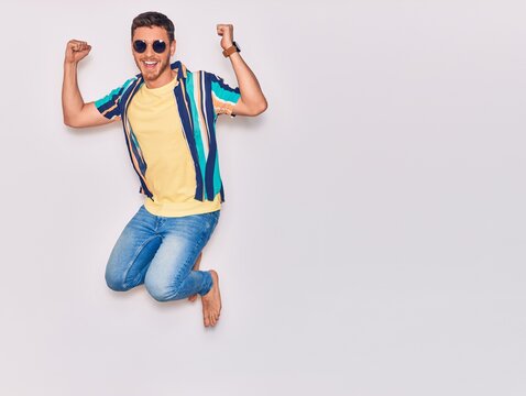 Young handsome hispanic man wearing casual clothes and sunglasses smiling happy. Jumping with smile on face celebrating with fists up over isolated white background