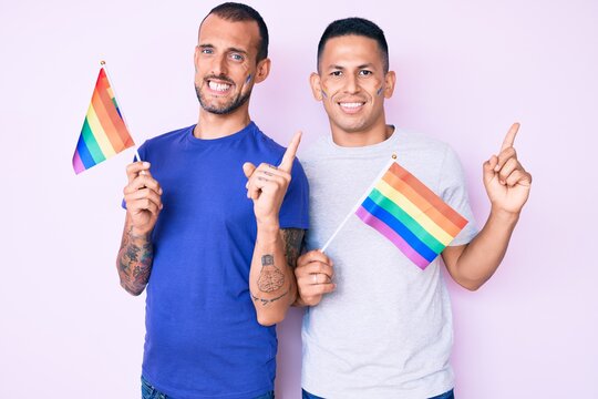 Young gay couple of two men holding rainbow lgbtq flags together smiling happy pointing with hand and finger to the side