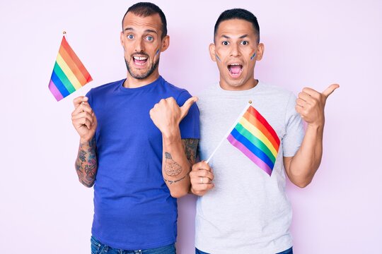 Young gay couple of two men holding rainbow lgbtq flags together pointing thumb up to the side smiling happy with open mouth