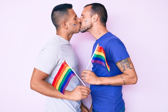 Young couple of gay men together in love holding homosexual pride colorful flag kissing with affection