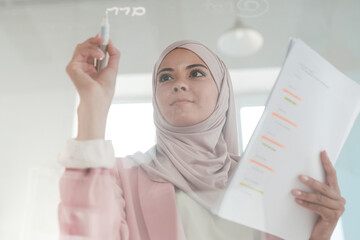 Young serious female broker in hijab holding financial papers and highlighter