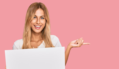 Naklejka premium Beautiful blonde young woman holding blank empty banner smiling happy pointing with hand and finger to the side