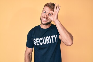 Young caucasian man wearing security t shirt doing ok gesture with hand smiling, eye looking through fingers with happy face.