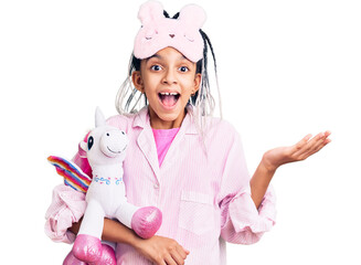 Cute african american girl wearing sleep mask and pajama holding pink teddy unicorn celebrating victory with happy smile and winner expression with raised hands
