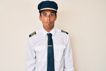 Young hispanic man wearing airplane pilot uniform making fish face with lips, crazy and comical gesture. funny expression.