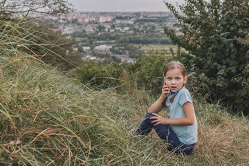 Naklejka na ściany i meble Little girl in a blue T-shirt talking on the phone. The child looks scared and speaks phone. Girl in the grass on a background of the city with a phone near her ear