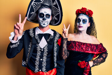 Young couple wearing mexican day of the dead costume over yellow smiling looking to the camera showing fingers doing victory sign. number two.
