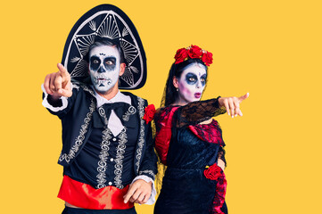 Young couple wearing mexican day of the dead costume over background pointing with finger surprised ahead, open mouth amazed expression, something on the front